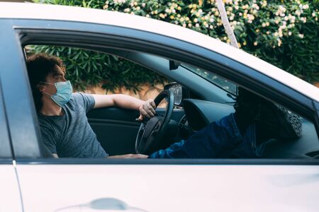 Young curly-haired man sits in car with his legs upstairs. Grey T-shirt and blue jeans. Resting after hard day. Guy in protective mask. Coronavirus epidemic. Boots with very pronounced rough solesの写真素材