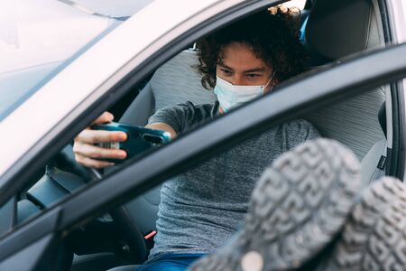 Young curly-haired man sits in car with his legs upstairs. Grey T-shirt and blue jeans. Takes selfie on mobile phone. Guy in protective mask. Coronavirus epidemicの写真素材
