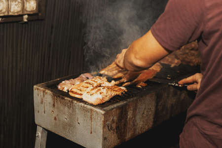 Hand of young man grilling some meat and fish on long rectangular barbecue with smoke on the street outdoor. Summer barbecue cooking. Barbecue with raw vegetables. BBQ on the terraceの写真素材