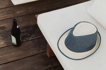 A blue and white hat lies on a white couch and a bottle of beer is blurred against a brown wooden floor near the pool. Party time on summer. Drinking alcohol. Mood. Postcardの写真素材