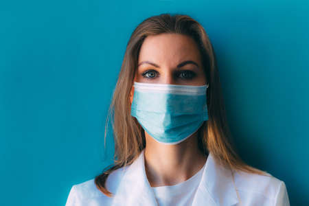 Portrait of young female doctor in medical mask and white gown on blue background. Female doctor in clinic in surgical mask with space for text. Head and shoulders portrait of intern in hospitalの写真素材