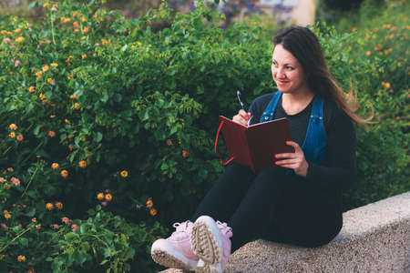 Young woman on bench in park with notebook and pen on green background. Wish Marathon. Writing wishes and dreams in a notebook. Life with a clean slate. Planning a new week. Life after quarantineの写真素材