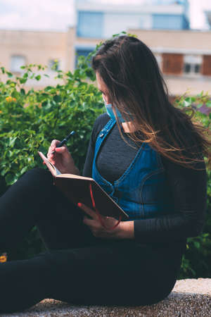 A young woman in a medical mask sits in a park and writes something in a notebook. Life after quarantine during the coronavirus epidemic pandemic. New reality. Respiratory protection. Green backgroundの写真素材