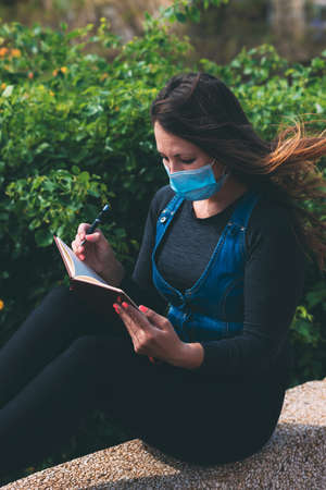 A young woman in a medical mask sits in a park and writes something in a notebook. Life after...