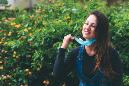 A young woman smiles and took off a blue medical mask and rejoices at the end of quarantine, in the park, outdoor. Life after quarantine during the coronavirus epidemic. New reality. Green backgroundの写真素材