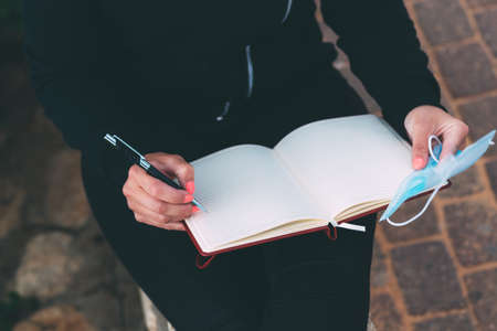 Side photo of blank notebook in female hands together with a pen and blue medical mask on left hand,...