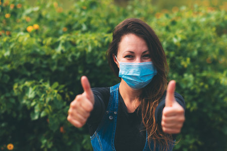 A young woman in a blue medical mask rejoices at the end of quarantine, shows with her hands a super...