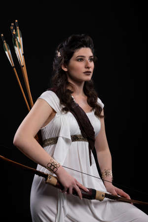 Portrait of a archer woman in white dress with bow and arrow on black background. Greek goddess Artemis. Studio photo shoot. Professional makeup and hairstyle for a brunette with long black hairの写真素材