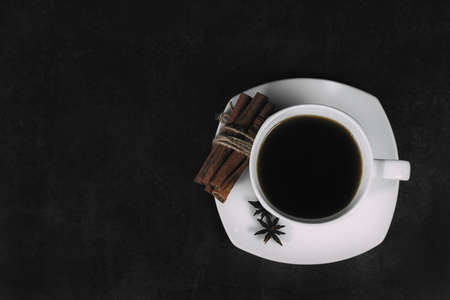 White cup with coffee and saucer, with anise star and cinnamon bunch, on dark textured background. A good idea for the menu or signboard of a restaurant or cafe, cafeteria. Top view, space for textの写真素材
