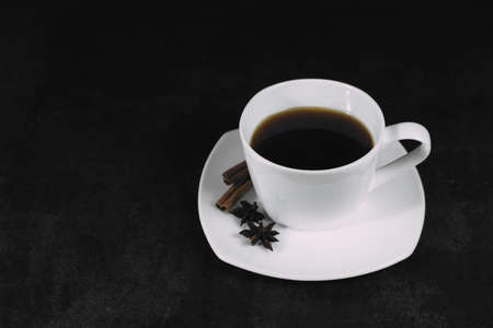 White cup with coffee and saucer, with anise star and cinnamon sticks, on dark textured background. A good idea for the menu or signboard of a restaurant or cafe, cafeteria. Side view, space for textの写真素材