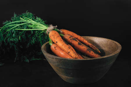 Close-up bouquet of fresh carrots with green leaves on wooden bowl on dark background. Organic eco food. Carrots with green tops tied with rope. Healthy diet. Freshly picked vegetables with groundの写真素材