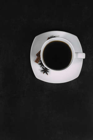White cup with coffee and saucer, with anise star and cinnamon sticks, on dark textured background. A good idea for the menu or signboard of a restaurant or cafe, cafeteria. Top view, space for textの写真素材