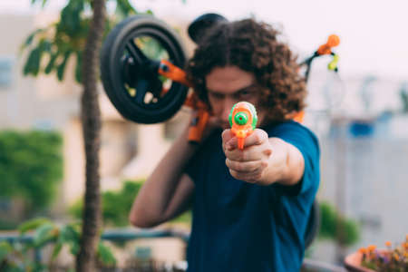 Blurred young man on Father's Day with water pistol and bicycle in hands, tired after playing with children, stands on the balcony in a blue T-shirt against the background of blurry palm treesの写真素材