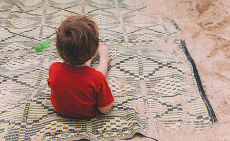 Father's day. Little son sits alone on the beach and waits for dad. Happy loving family. A lonely child in red T-shirt sits on a bedspread from the back. Before you go swimming in the seaの写真素材