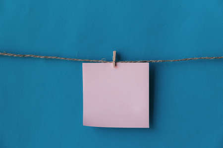 One pink sheet of paper with place for text hanging on rope isolated blue background. To do list. Pink sticky notes with cloth pegs. Pink notes paper. Reminder.の写真素材