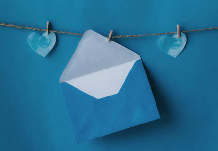 One opened blue envelope hanging on rope isolated blue background with two blue hearts on sides. Write a letter to a friend. Postal envelope. Gift envelope for postcards.の写真素材
