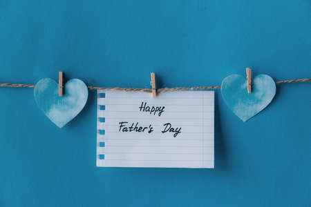 White notebook in line paper greeting card with black handwritten lettering happy fathers day hanging on brown rope with clothespins isolated on blue background with two blue hearts on the sidesの写真素材