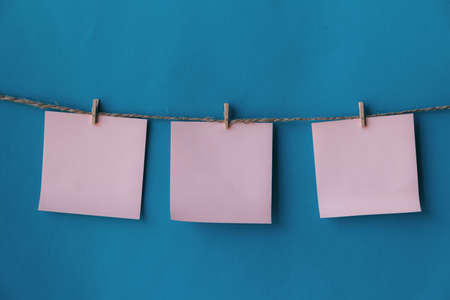 Three pink sheets of paper with place for text hanging on rope isolated blue background. To do list. Pink sticky notes with cloth pegs. Pink notes paper. Reminder.の写真素材