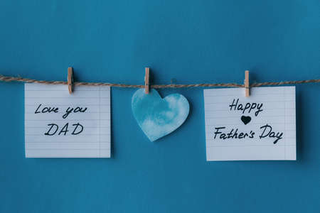 Two white notebook in line paper greeting card with black handwritten lettering happy fathers day and I love you dad hanging on brown rope with clothespins isolated on blue background with blue heartの写真素材