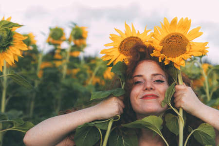 Beautiful young girl with red wavy hair and freckles enjoying nature on the field of sunflowers. The concept of summer and sun. Sunflower season in Israel. Sunflowers instead of ears.の写真素材