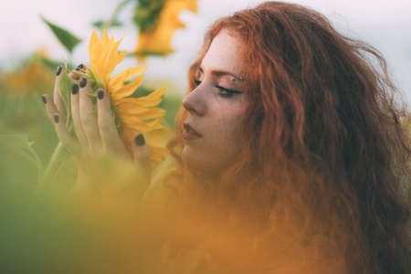 Close-up portrait of beautiful young girl with red wavy hair and freckles enjoying nature on the field of sunflowers. The concept of summer and sun. Sunflower season. Holding sunflower on her handsの写真素材