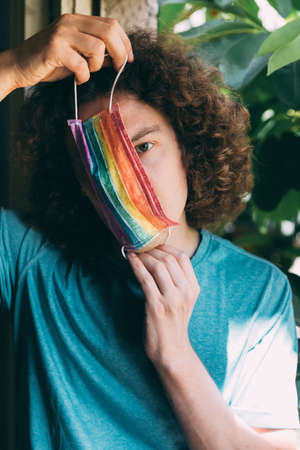 Man in a rainbow medical mask. Young guy with long curly hair wearing painted surgical mask on face. Pride protection. Lgbt medicine mask. Pride concept. Period of COVID-19 coronavirus. Close-upの写真素材