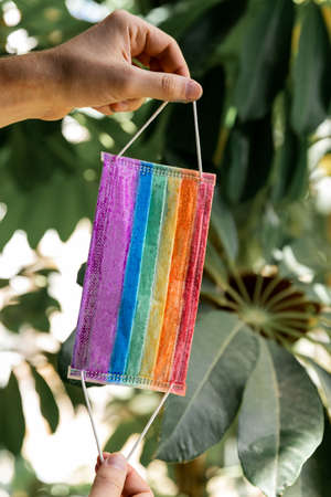 Pride. Medical LGBT mask in rainbow colors in male hands on a blurred background of green leaves. Covid 19 protection. LGBT community flag. Protective medical face mask. Stay home concept. Bannerの写真素材