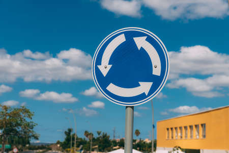 Close-up view of blue and white roundabout sign on the street with grass, trees, road and blue sky...