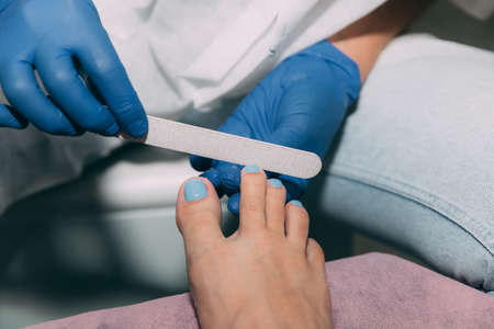 Pedicure in salon. Foot care treatment and nail. File filing with a nail file after applying gel polish. Master in blue gloves makes pedicure with manicure machine. Concept of beauty care and healthの写真素材