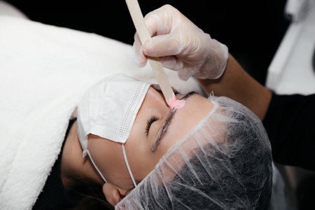 Facial hair removal procedure by hot wax on wooden stick, preparation before permanent makeup tattoo on eyebrows. Safe procedure using medical masks and sterile gloves. New reality in beauty worldの写真素材