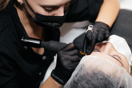 Professional beautician master of permanent makeup in black medical mask and gloves makes tattooing eyebrows. Concept of beauty salon and permanent make-up. New reality in beauty world. Covid 19の写真素材