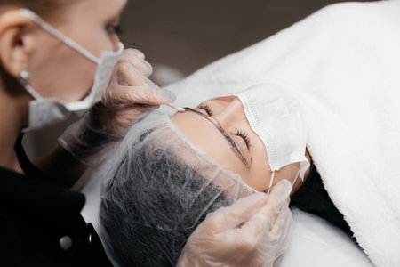 Removing anesthetic cream from the eyebrows before the permanent makeup procedure. Master and client in medical masks and gloves. Permanent makeup eyebrows conturing. New reality in beauty worldの写真素材
