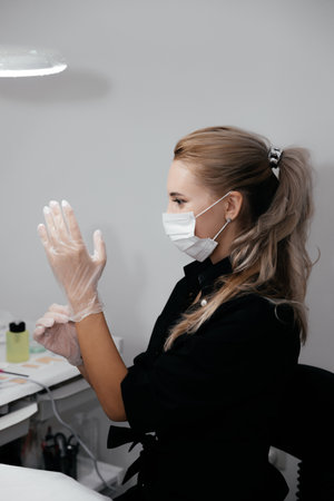 Young permanent make up master in surgical mask wearing gloves and prepares to receive clients in beauty salon. Prevention coronavirus Covid-19 on quarantine period. Ð¡osmetologist, beautician in maskの写真素材