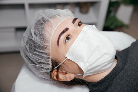 Happy client in medical mask after permanent makeup eyebrows. Woman with makeup lying in the sterile cap on a procedure of microblading with finished eyebrows. Beauty salon. Result of tattoo.の写真素材