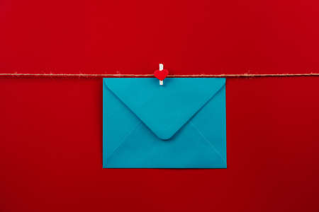 One closed blue envelope with a clothespin with a heart hanging on rope isolated red background. Write a letter to a friend. Postal envelope. Gift envelope for postcards. Happy Valentines day, 14thの写真素材