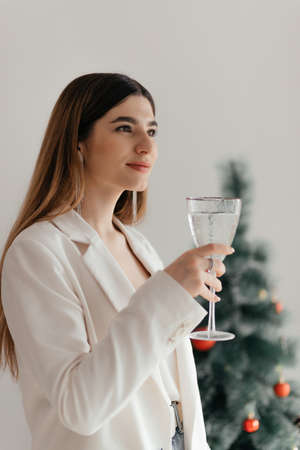 A young pretty girl in a white jacket and earrings with brown long hair smiles with a glass of champagne against the background of a festive Christmas tree and a white wallの写真素材