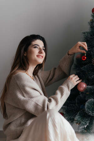 Young beautiful brunette wearing brown pullover decorates the Christmas tree with toys and is in...