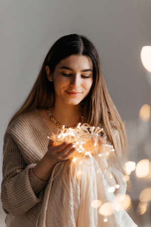 Pretty brunette wearing a beige sweater posing with gold garland in her hands, and big bokeh in the foreground. She is looking at garland and she is happy. New year and Christmas conceptの写真素材