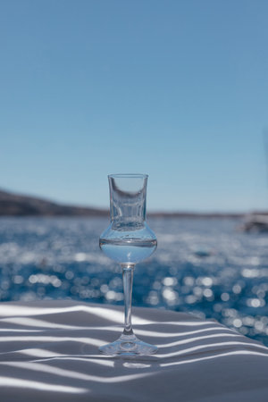 Closeup view of one fantastic beautiful glass with transparent national alcohol drink on white table with play of shadows in Santorini, Greece, with blurred Aegean sea with bokeh on backgroundの写真素材