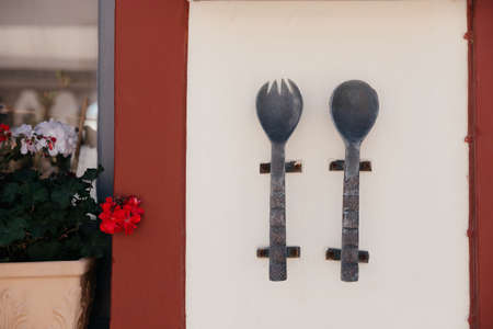 Large metal and old spoon and fork with carvings and patterns on them hang on the wall of cafe or restaurant on Santorini island, Oia, Greece. Signboard on the street near window and flower in potの写真素材