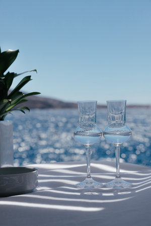 Closeup view of two beautiful glasses with transparent national alcohol drink on white table with play of shadows and green flower in Santorini, Greece, with blurred Aegean sea and ship on backgroundの写真素材