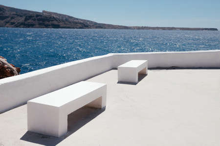 Two white benches on the coast of Santorini and the sea with blue sky and mountains in the...