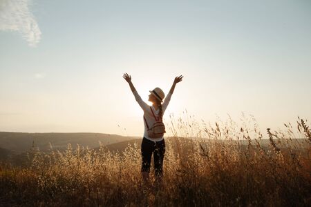 A young girl in a hat and a backpack stands on top of the mountain and raised her hands to the skyの写真素材