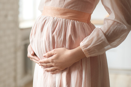 Young pregnant woman at home maternity concept wearing dress touching belly close-upの写真素材