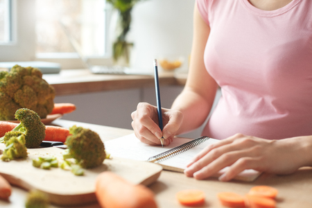 Young pregnant woman in the kitchen at home writing shopping list close-upの写真素材