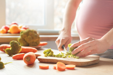 Young brunette woman expecting baby standing in the kitchen preparing salad cutting broccoli close-up near windowの写真素材