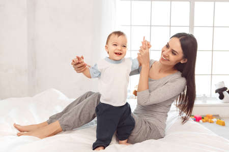 Motherhood. Mother helping son to walk sitting on bed smiling cheerfulの写真素材