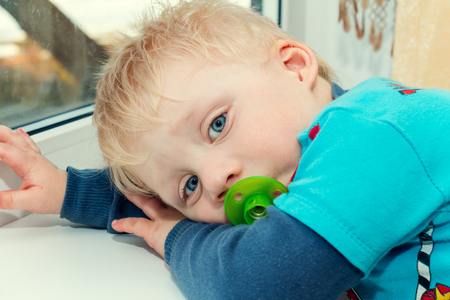 Little funny boy with a pacifier is sitting near the window.の写真素材