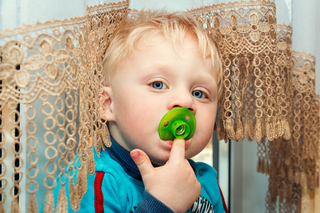 Little funny boy with a pacifier is sitting near the window.の写真素材