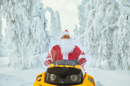 Authentic Santa Claus is riding a snowmobile through the winter forest. Magnificent snow-covered landscape.の写真素材